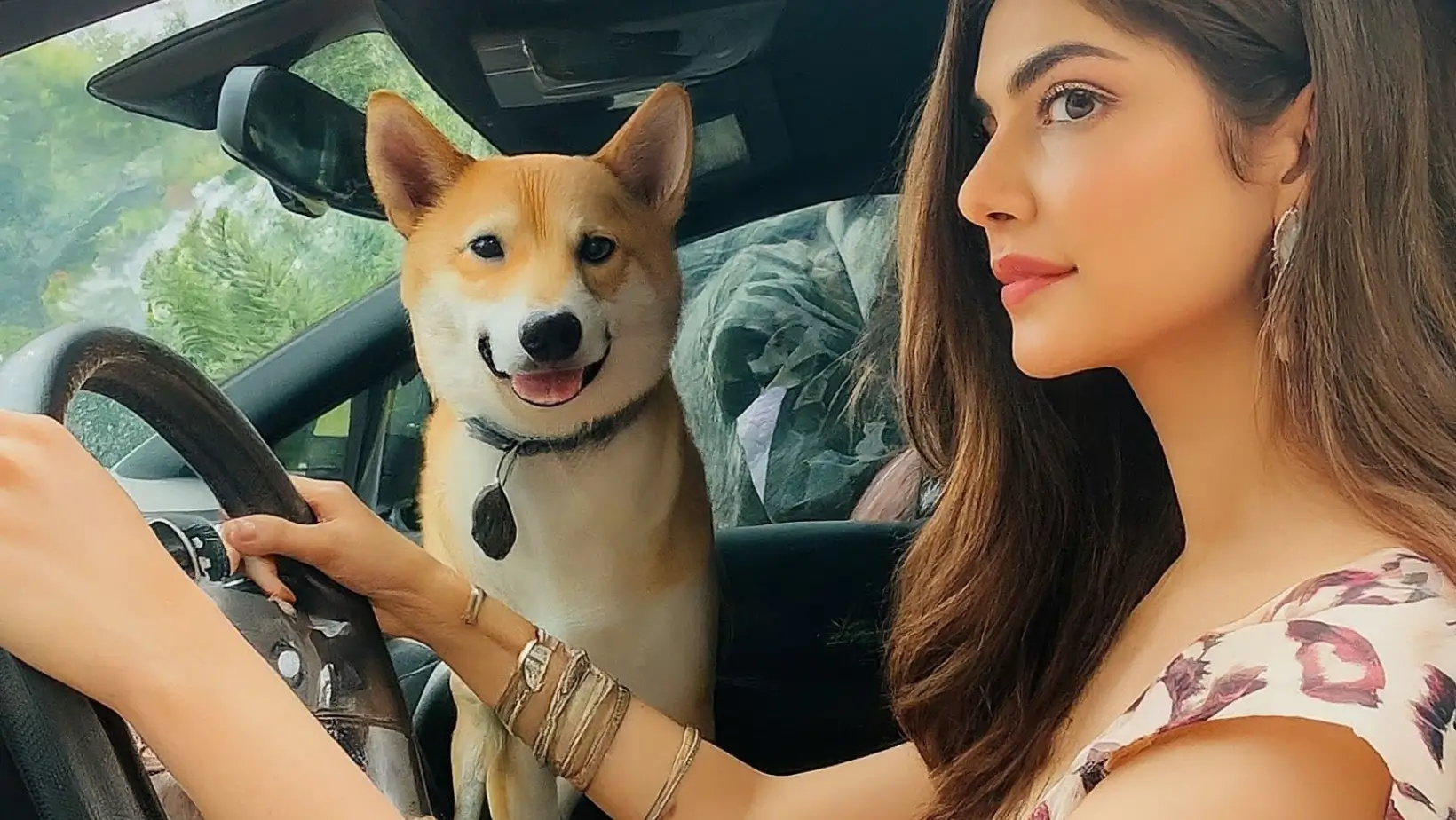 a beautiful lady is driving the car and a dog is stand on front seat